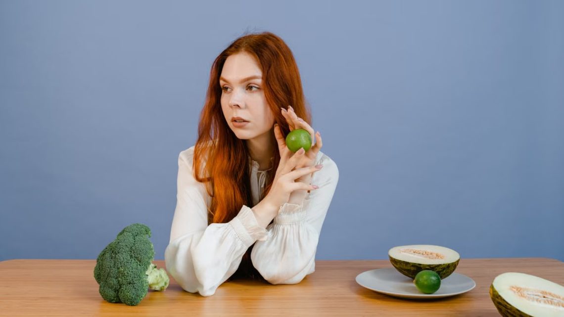 Caucasian woman sitting at table holding lime with broccoli and melon.